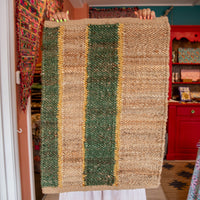 Person holding a woven rug with green, brown, and gold stripes in a room with colorful decor.