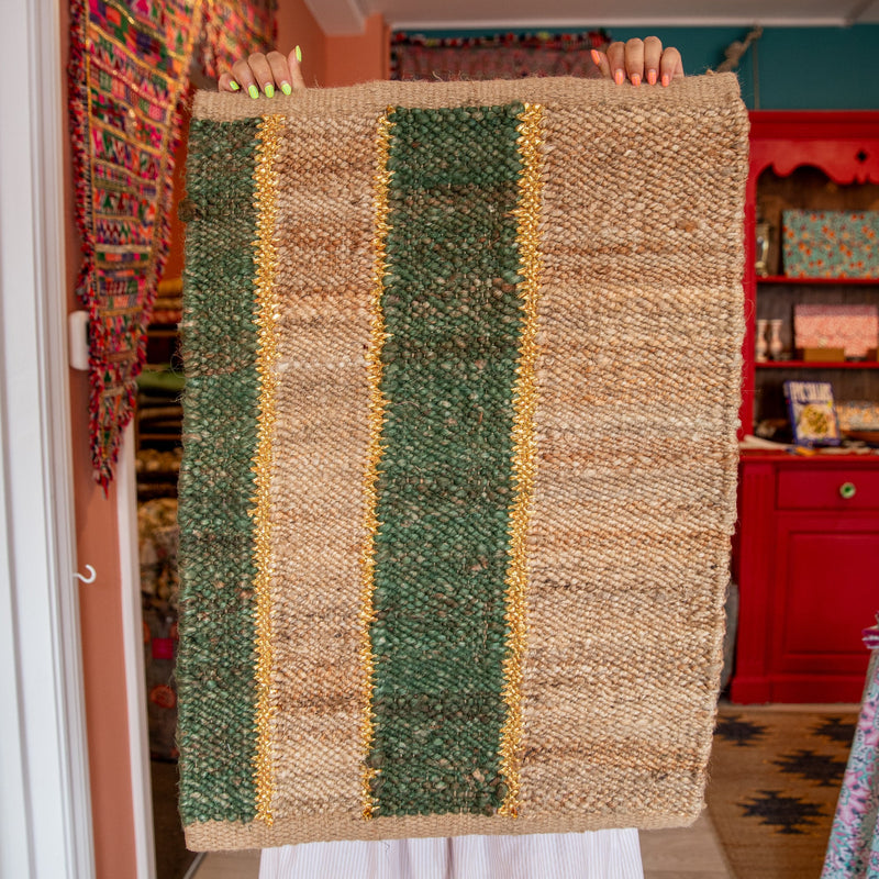 Person holding a woven rug with green, brown, and gold stripes in a room with colorful decor.