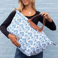 A woman holds a large tote bag with a blue floral block print on white fabric and brown leather handles, standing against a light blue brick wall.