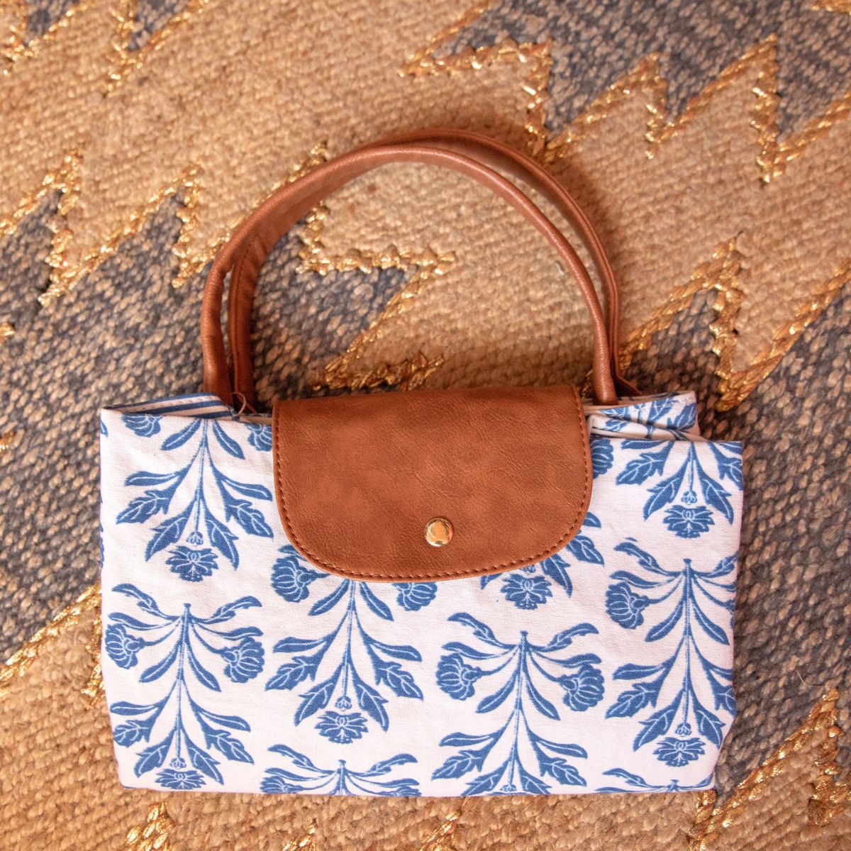 A structured handbag with a white canvas body featuring a blue floral damask pattern, a brown faux-leather flap closure with a gold snap button, and a brown top handle, laid flat on a textured patterned rug.