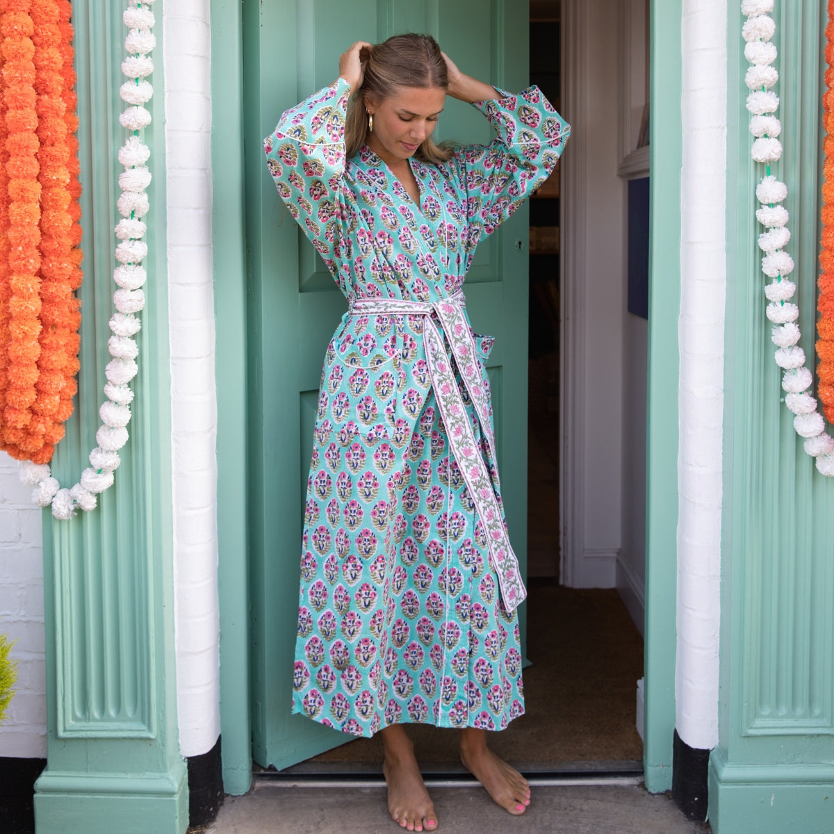 A woman wearing a long-sleeved, tie-waist floral print robe stands barefoot in a doorway. The robe has a light aqua background with a pink and purple floral pattern.