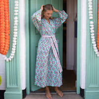 A woman wearing a long-sleeved, tie-waist floral print robe stands barefoot in a doorway. The robe has a light aqua background with a pink and purple floral pattern.