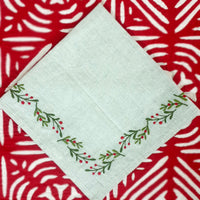 A linen napkin with embroidered holly berries design on a neutral background, displayed on a patterned surface.
