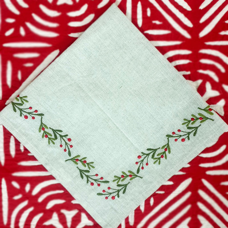 A linen napkin with embroidered holly berries design on a neutral background, displayed on a patterned surface.