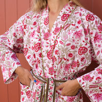 A close-up of a woman wearing a white cotton robe with a vibrant pink and red floral block print, featuring green leafy accents and a matching tie belt.