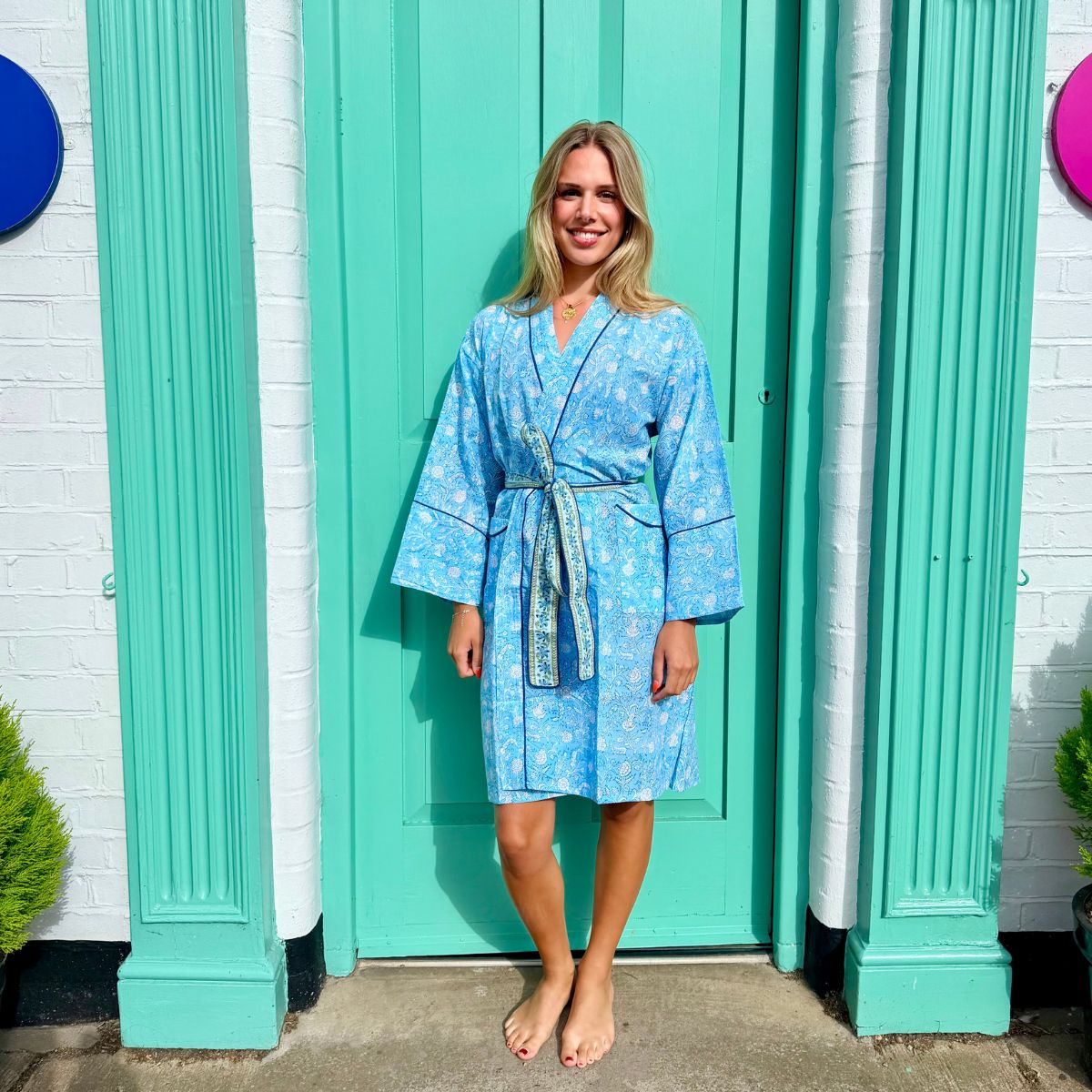 A woman stands barefoot in front of a bright turquoise door wearing a light blue patterned robe with a matching tie belt. The background includes white brick walls and decorative columns.
