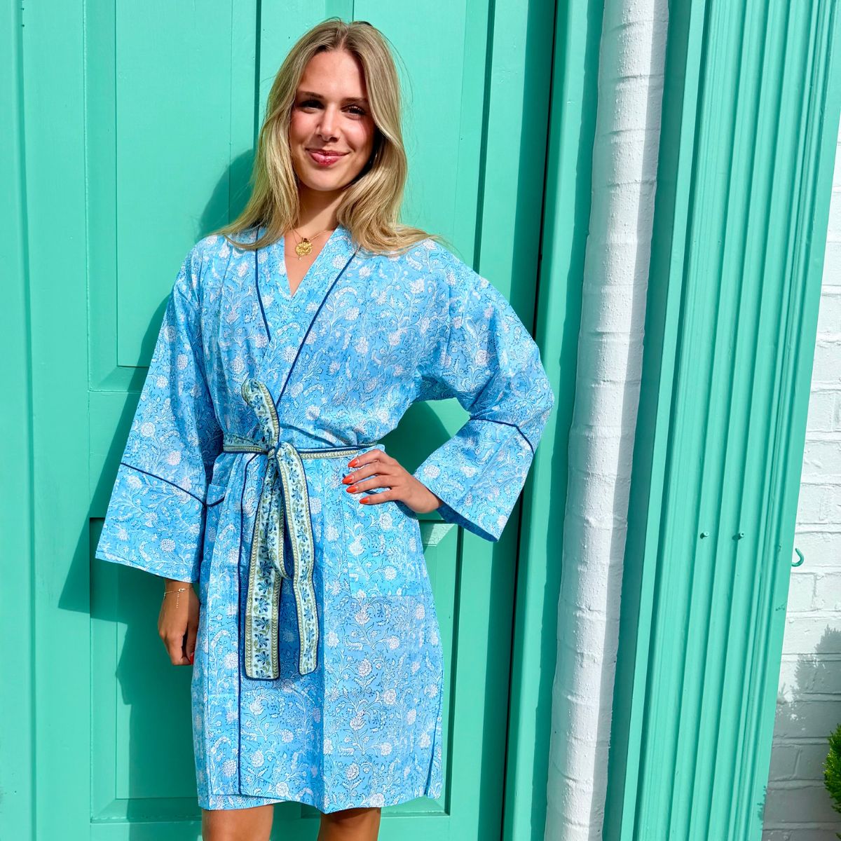 A woman wearing a light blue floral print bathrobe with a matching tie belt, standing in front of a turquoise door.
