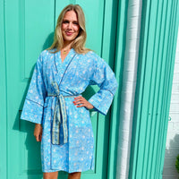 A woman wearing a light blue floral print bathrobe with a matching tie belt, standing in front of a turquoise door.