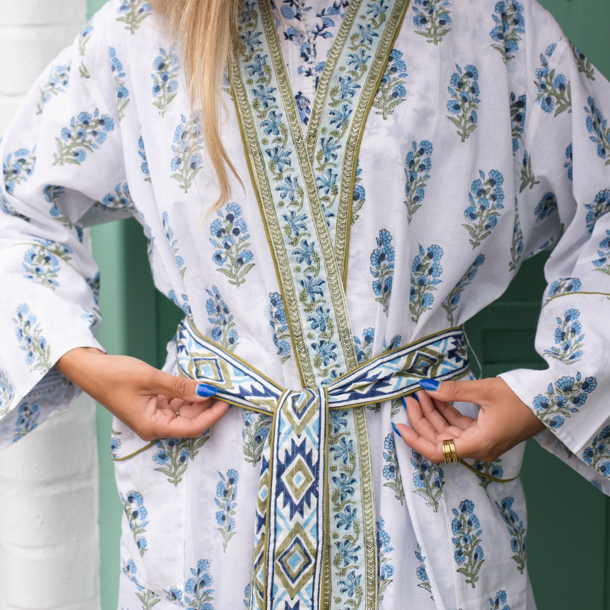 A close-up of a person wearing a white kaftan robe with blue floral prints and green borders, tying a matching patterned belt. 