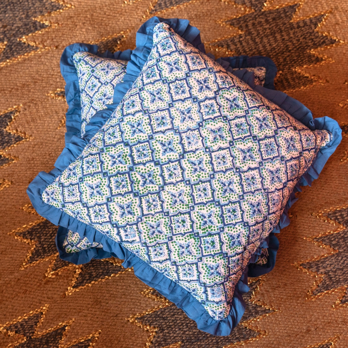A square decorative throw pillow with a blue ruffled border and a white, blue, and green geometric patterned fabric, displayed on a brown patterned rug.