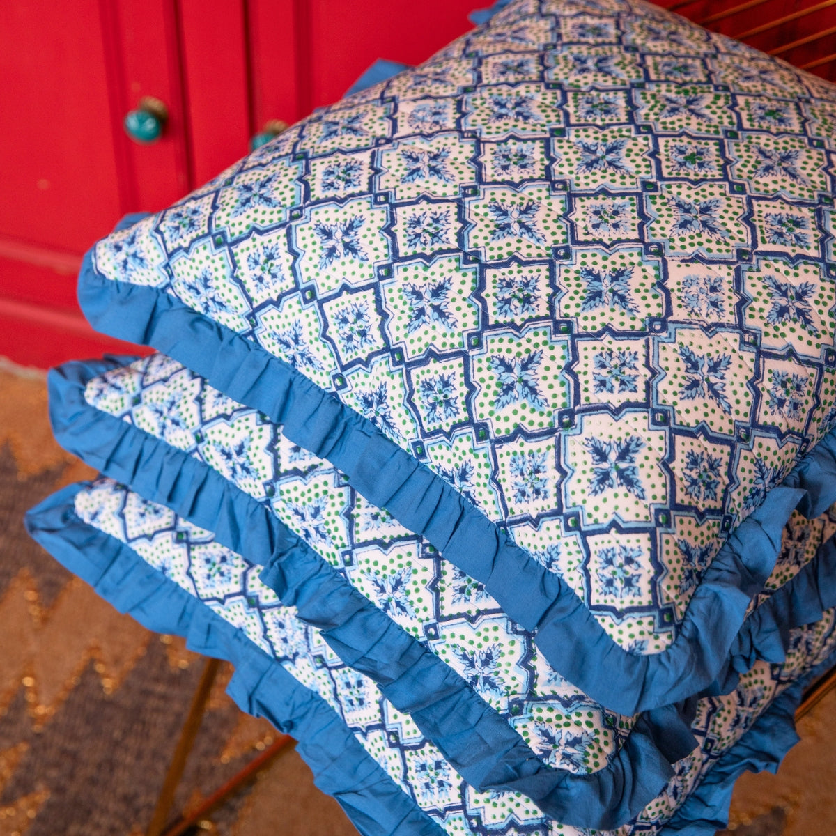 Three decorative throw pillows with blue ruffled edges and a blue, white, and green geometric tile pattern, stacked on a surface with a red cabinet in the background.