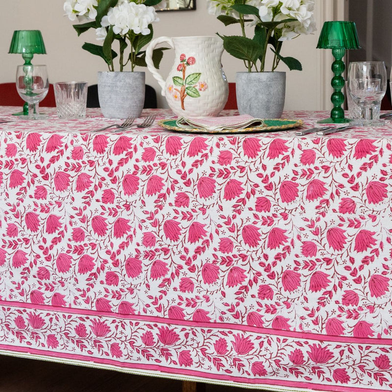 A white tablecloth with a vibrant pink and green floral pattern, styled on a dining table with green candlesticks, glassware, and white flowers in gray pots.