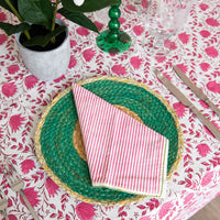 A styled table setting featuring a round green woven charger plate with a gold braided rim, topped with a pink and white striped napkin. The table is covered in a white tablecloth with a vibrant pink floral pattern, accompanied by silver cutlery, a green glass candlestick, and a potted plant.