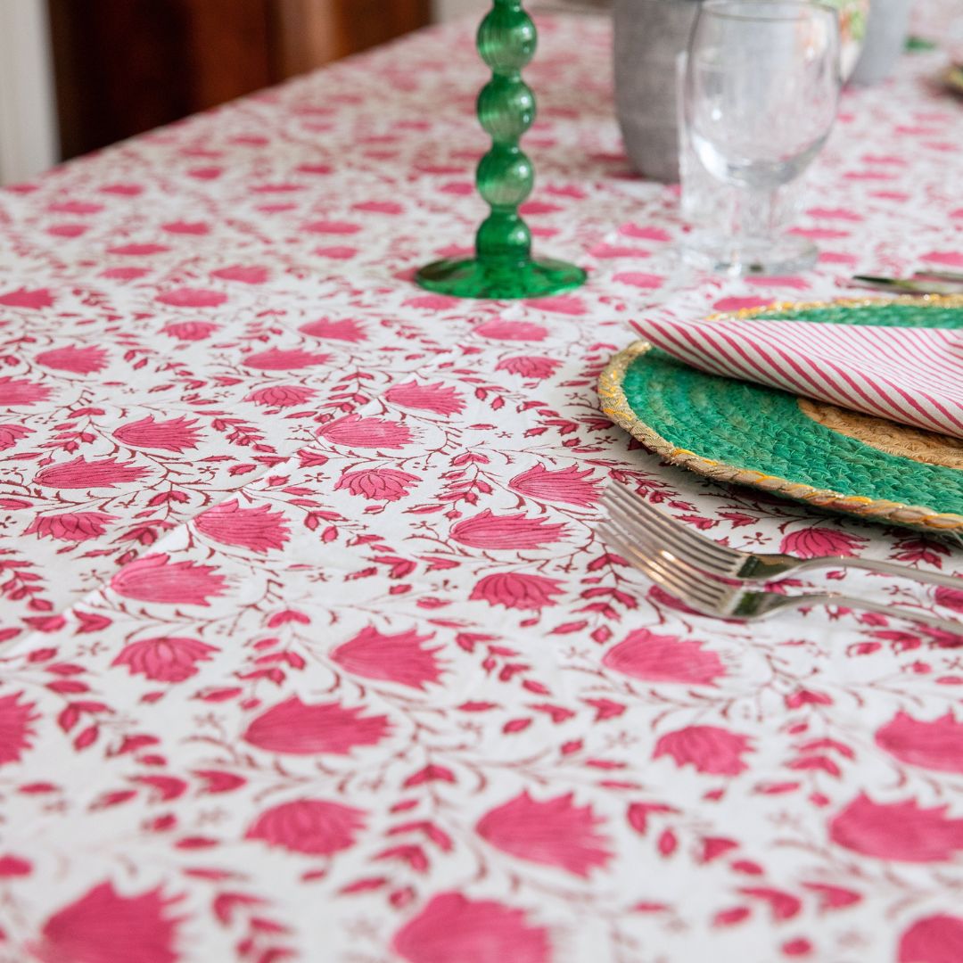 A close-up of a white tablecloth with a vibrant pink floral block print pattern, styled with a green glass candlestick, a woven green placemat, and a striped napkin on a dining table.