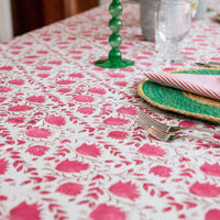 A close-up of a white tablecloth with a vibrant pink floral block print pattern, styled with a green glass candlestick, a woven green placemat, and a striped napkin on a dining table.