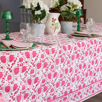 A white tablecloth with a vibrant pink floral block print pattern, styled on a dining table with pink striped napkins, green glass candle holders, and white flower arrangements.