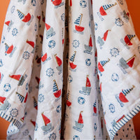 A white quilted baby blanket featuring a colorful nautical print of red, gray, and blue sailboats, life buoys, and ship's wheels, draped against an orange background.
