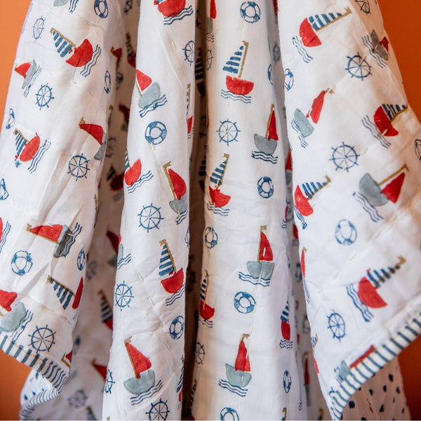 A white quilted baby blanket featuring a colorful nautical print of red, gray, and blue sailboats, life buoys, and ship's wheels, draped against an orange background.