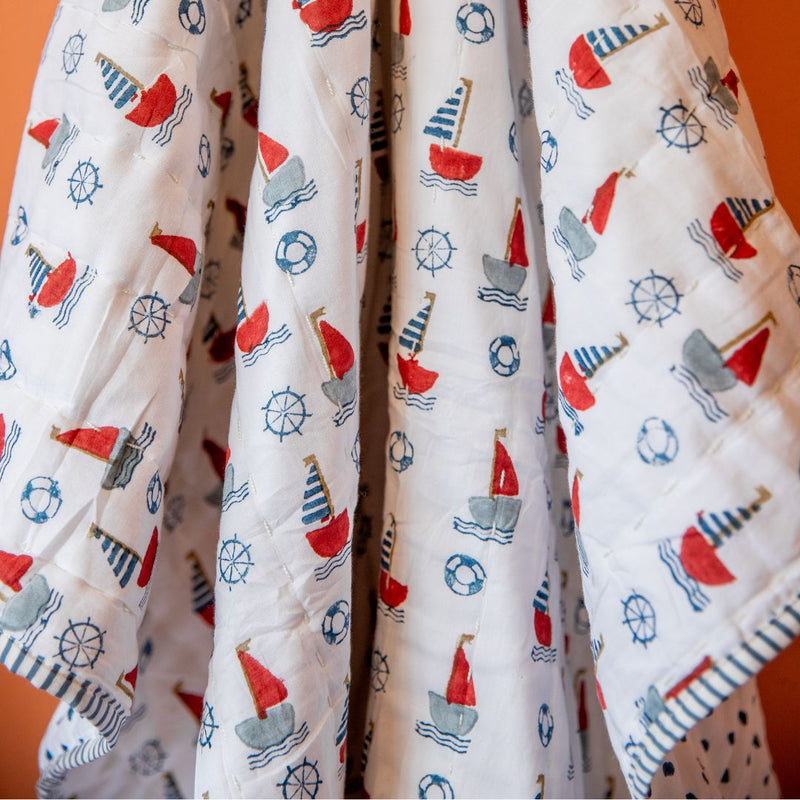 A white quilted baby blanket featuring a colorful nautical print of red, gray, and blue sailboats, life buoys, and ship's wheels, draped against an orange background.