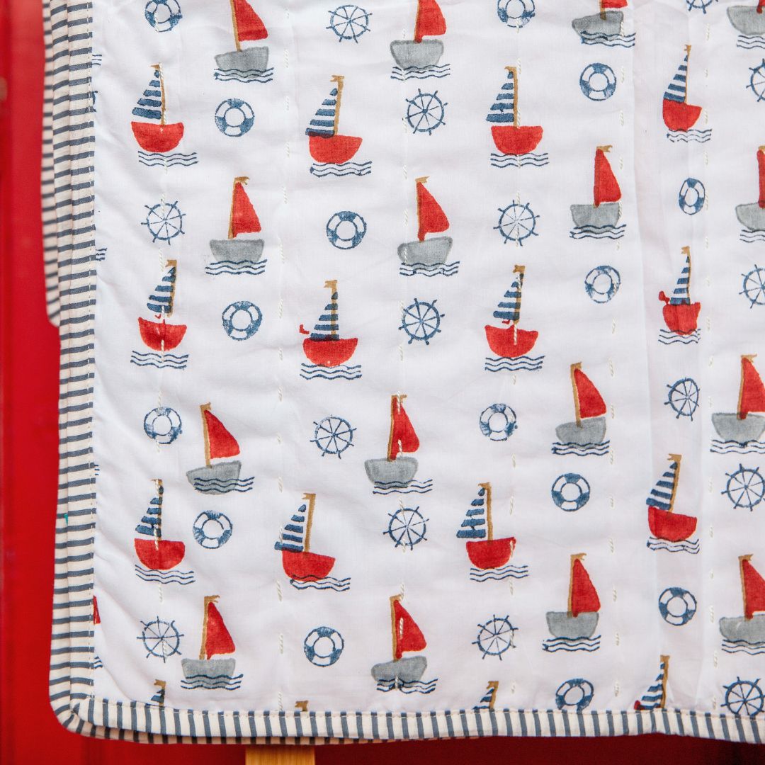 A close-up of a quilted baby blanket featuring a nautical sailboat pattern on a white background, with red and gray boats, blue wheels, and life preservers. The blanket has a striped border and is shown folded over on a red surface.