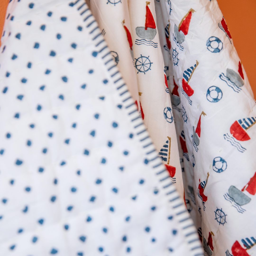Close-up of folded baby blankets or quilts showing three different nautical-themed prints: a white quilt with small blue hearts, a cream quilt with red and gray sailboats, and a white quilt with blue life preserver and sailboat motifs.