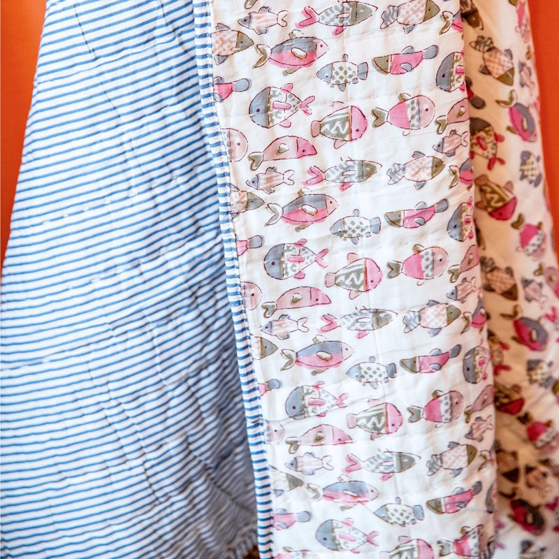 Close-up of a quilted blanket or comforter showing three fabric panels: a blue and white striped panel, a white panel with a pink, gray, and blue fish pattern, and a cream panel with a red and pink fish pattern, set against a solid orange background.