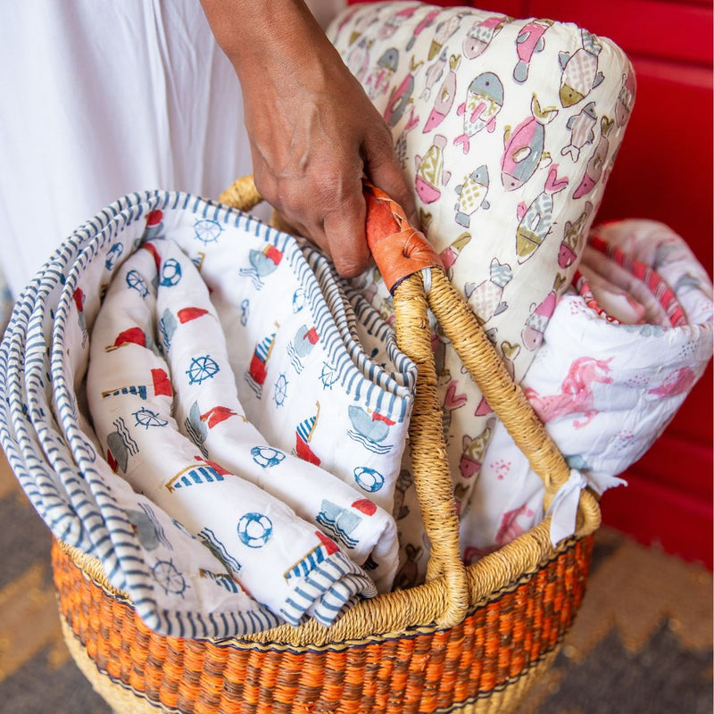 A woven basket filled with folded baby blankets and swaddle fabrics in nautical and fish patterns, held by a person wearing a white top. The fabrics are white with blue, red, pink, and yellow printed motifs.
