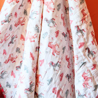 A close-up of a white quilted blanket or comforter featuring a whimsical print of pink, red, and gray horses and small hearts, draped against a solid orange background.