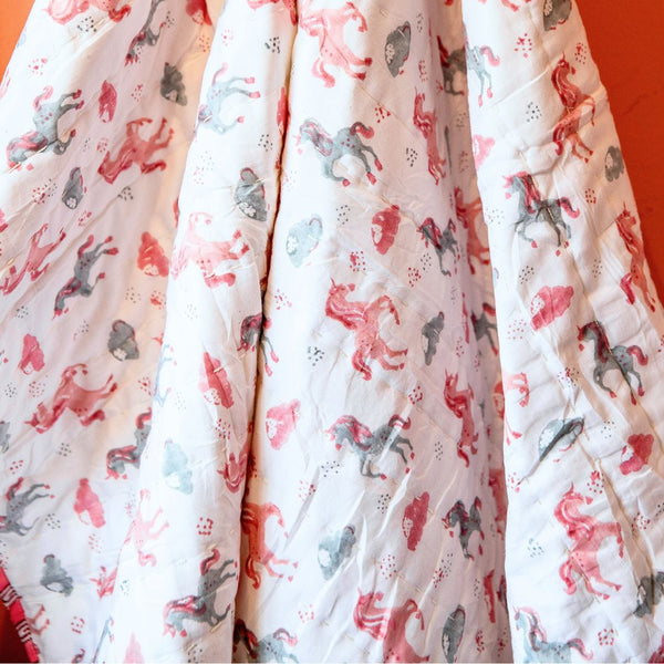 A close-up of a white quilted blanket or comforter featuring a whimsical print of pink, red, and gray horses and small hearts, draped against a solid orange background.
