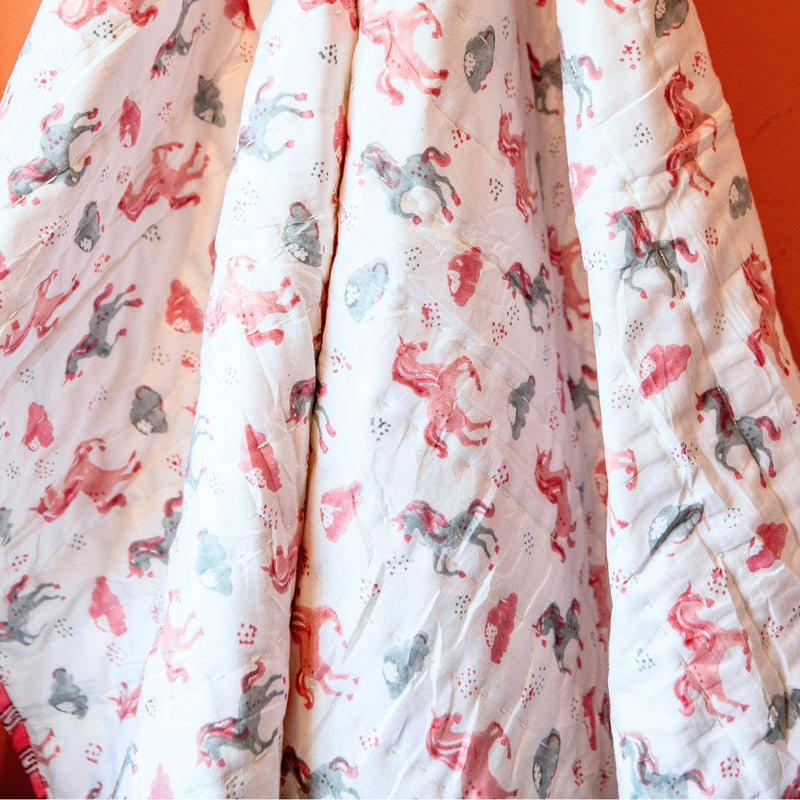 A close-up of a white quilted blanket or comforter featuring a whimsical print of pink, red, and gray horses and small hearts, draped against a solid orange background.