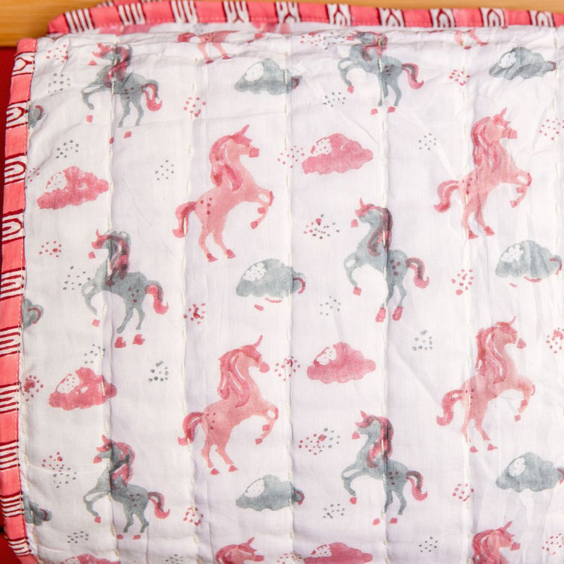 A close-up of a quilted blanket or comforter with a white background, featuring a repeating pattern of pink and gray cartoon horses and small red and gray dots. The blanket has a pink and white patterned ruffled trim along the left and bottom edges.
