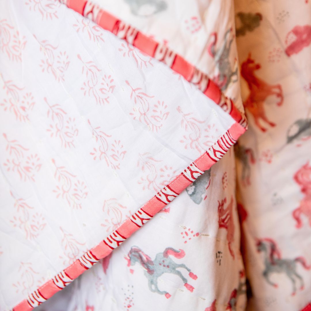 A close-up of a quilted baby blanket featuring a white background with a pink and gray unicorn pattern, accented by a pink and white striped border.