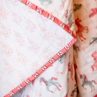 A close-up of a quilted baby blanket featuring a white background with a pink and gray unicorn pattern, accented by a pink and white striped border.